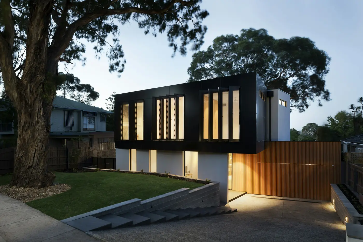 Night House: a black box with vertical lit windows above a white base, at dusk